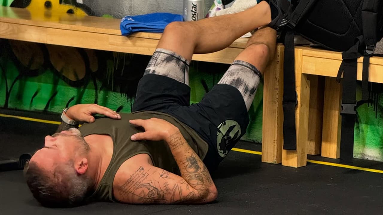 Tätowierter Athlet liegt erschöpft auf dem Fitnessstudio-Boden mit erhöhten Beinen auf einer Bank zur Erholung