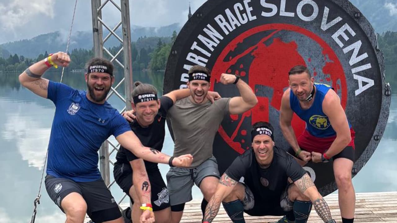 Fünf Athleten feiern gemeinsam nach dem Spartan Race in Slowenien vor dem offiziellen Rennschild am Bleder See