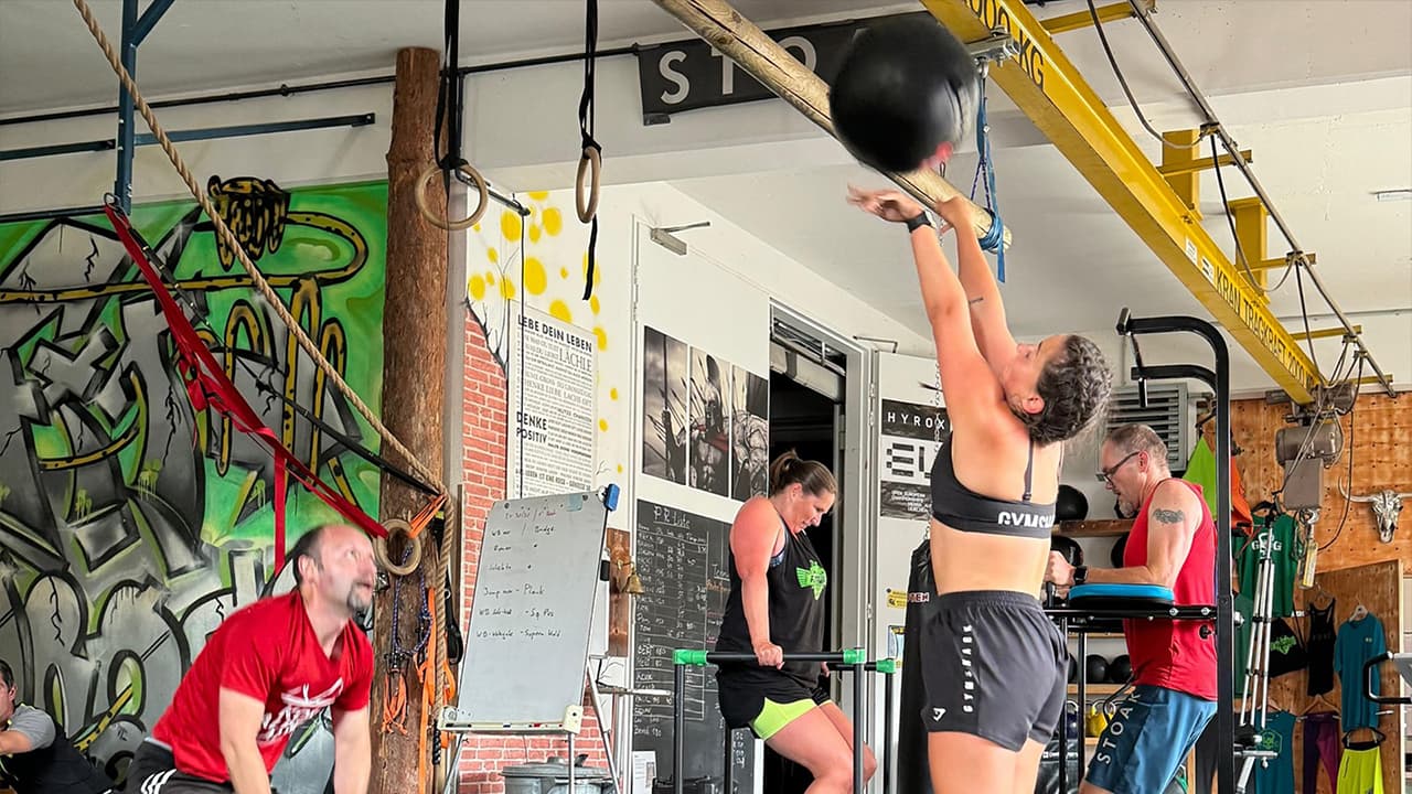 Athletin wirft Medizinball während intensivem CrossFit Training im Fitnessstudio mit anderen Sportlern